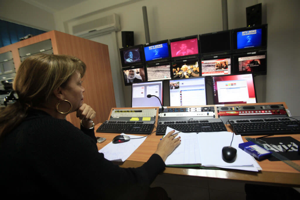 A producer works at a broadcast control desk in front of a wall of monitors.