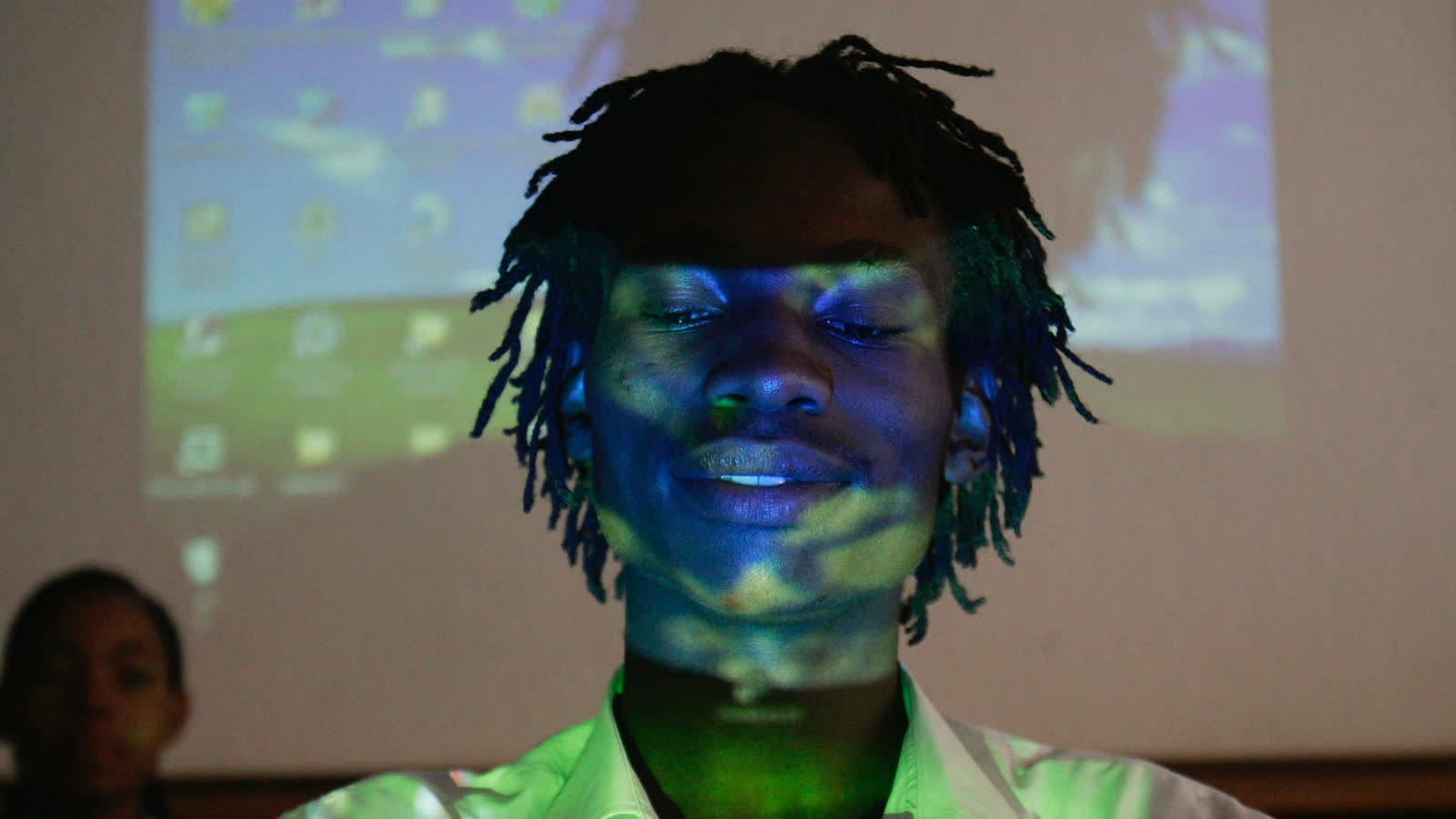 A young man stands in front of a projected computer screen, light falling across his face.
